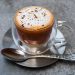 Rich aroma cappuccino in a clear glass mug with frothy foam and sprinkled chocolate on a metallic saucer.